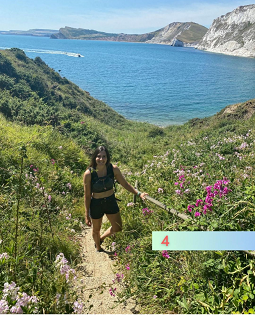 A white female with long brown hair smiles at the camera on a coastal path. 